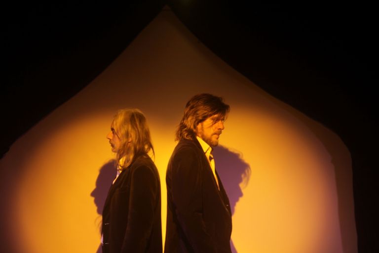 Musicians Matt Walker and Tex Perkins stand back-to-back in side profile under warm yellow spotlight in a moody portrait.